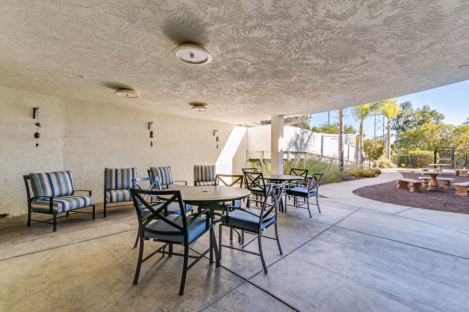 Covered outdoor seating area with several metal tables and chairs with cushions, adjacent to a garden area with stone benches and trees under a clear sky.