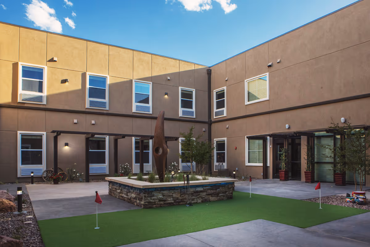 Courtyard of a two-story senior living facility with a central planter and sculpture, small putting green, and surrounding windows and entrances.
