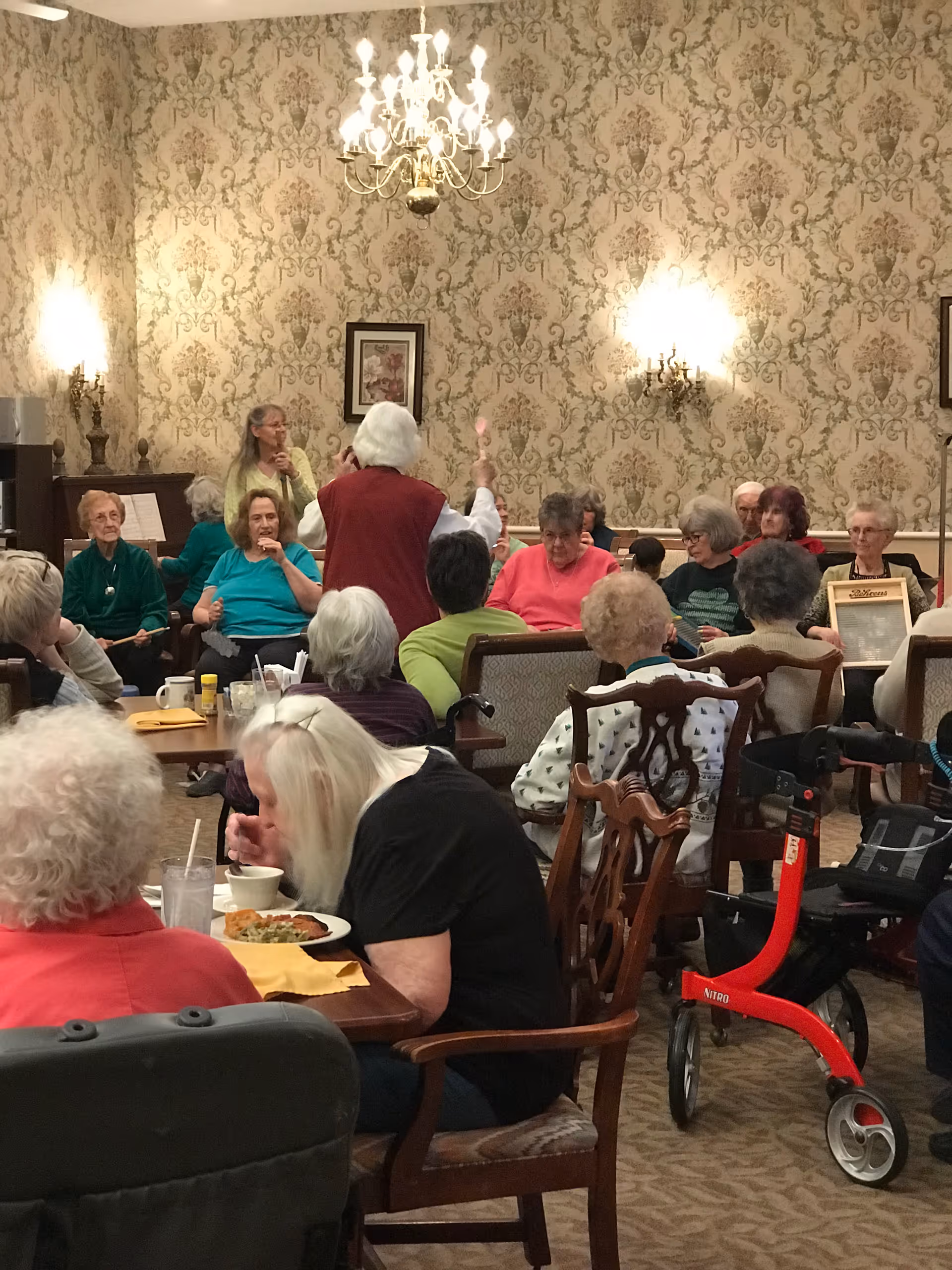 A group of elderly people gathered in a room with ornate wallpaper and a chandelier. Some are seated at tables eating, while others are seated in a semi-circle, seemingly engaged in an activity led by a woman standing in the center. A red walker is visible near the foreground.