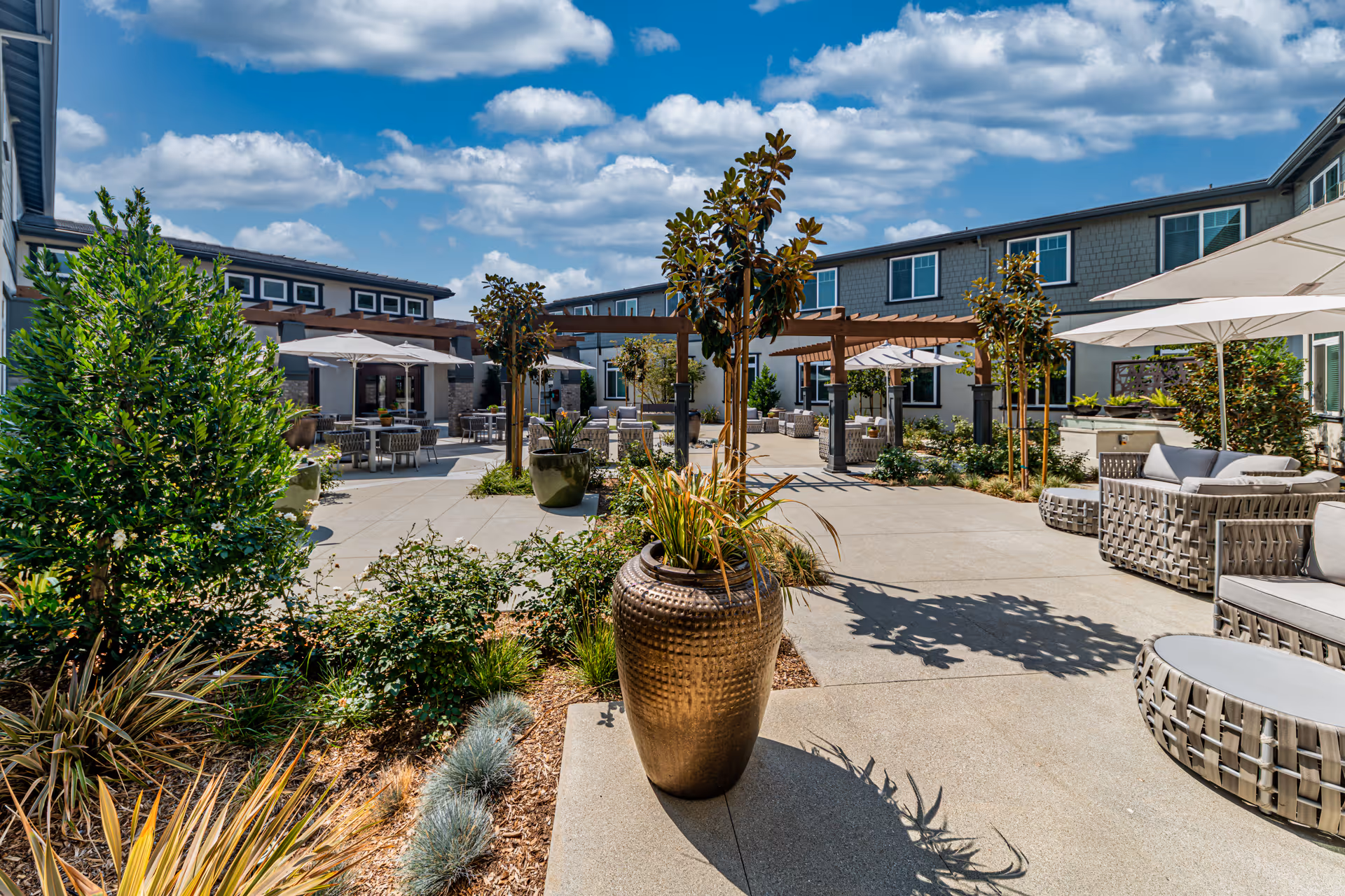 Outdoor courtyard area at Allara Senior Living featuring paved walkways, large decorative planters with plants, several small trees, patio furniture including cushioned chairs and tables with umbrellas, and a pergola structure under a partly cloudy blue sky.