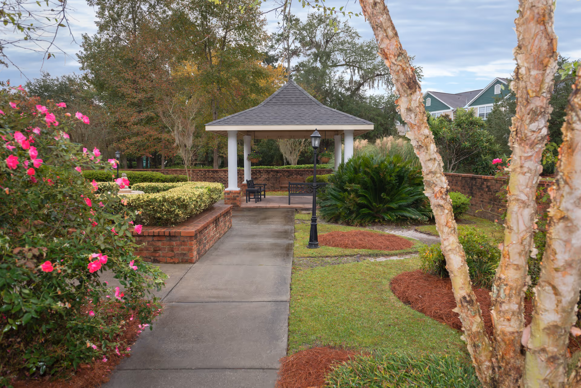 A paved walkway leading to a gazebo with white columns and a gray roof in a landscaped garden area. The garden features trimmed bushes, pink flowers, a black lamppost, and trees with autumn foliage. Residential buildings are visible in the background under a partly cloudy sky.
