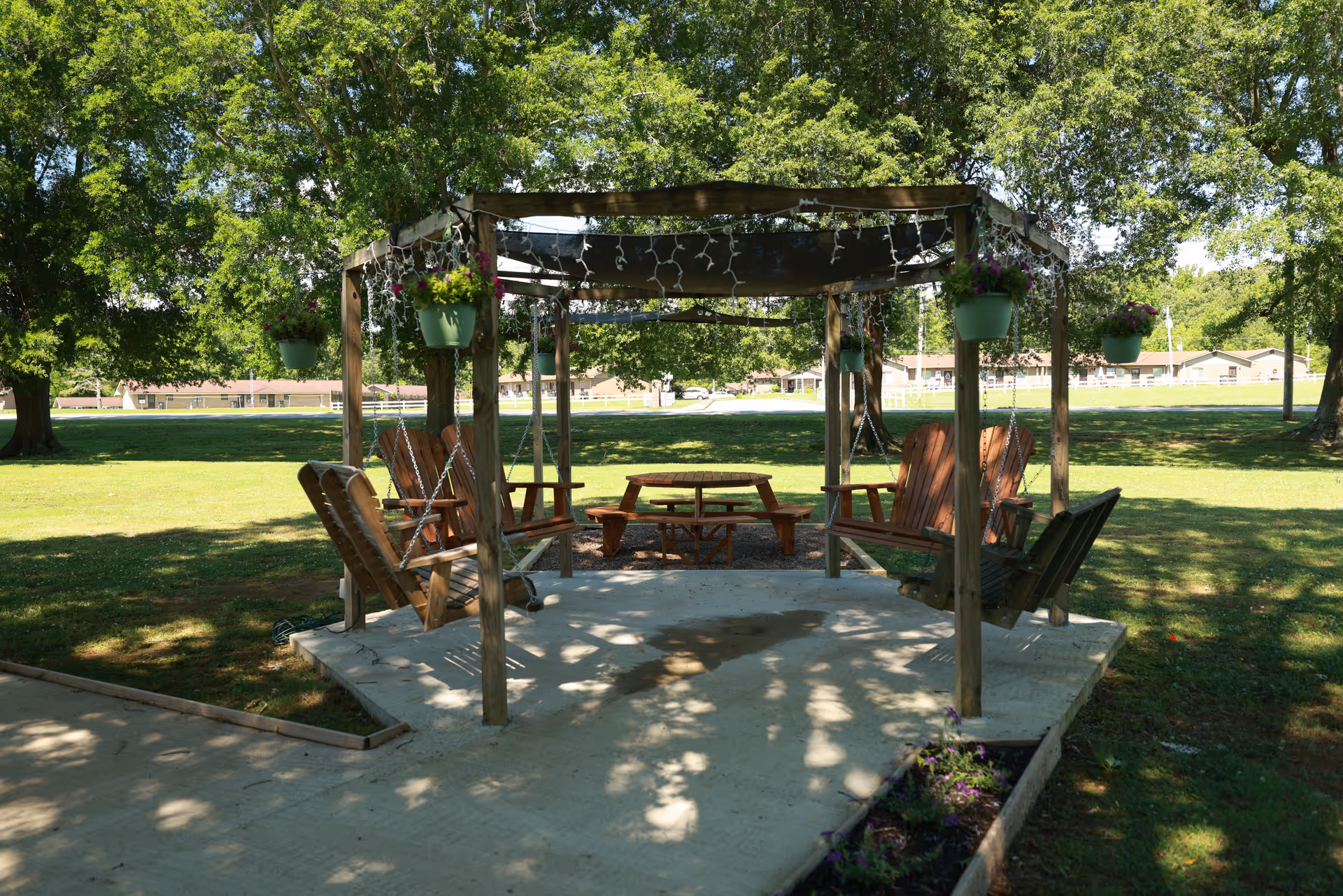 Wooden pergola with hanging flower pots and swing chairs arranged around a small picnic table on a shaded lawn.