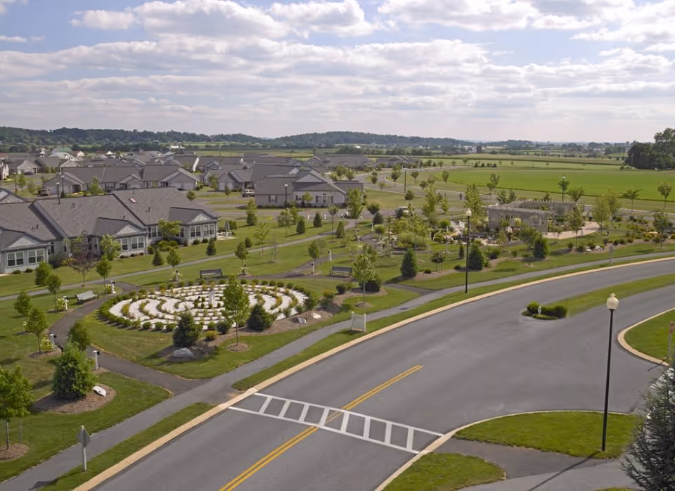 Aerial view of Garden Spot Village showing a residential community with single-story houses, landscaped gardens, walking paths, and a road with street lamps. The area is surrounded by green fields and trees under a partly cloudy sky.