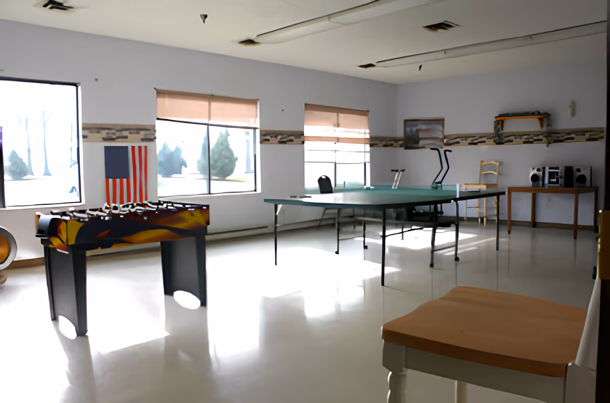 A bright recreational room with large windows letting in natural light. The room contains a foosball table, a ping pong table, several chairs, a small table with a stereo system, and an American flag hanging on the wall.