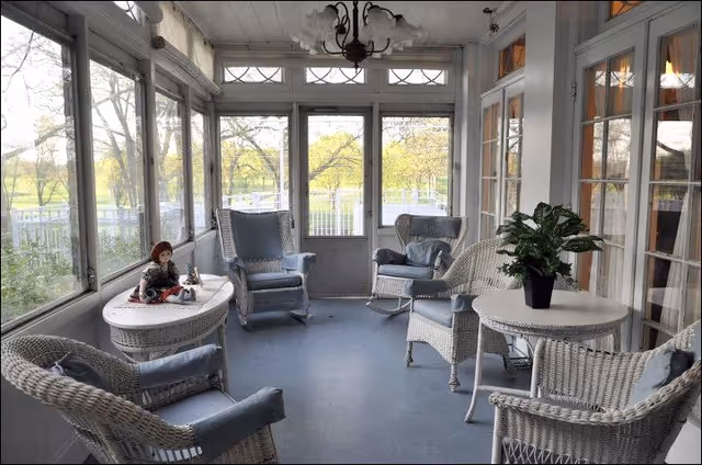 A bright enclosed porch with large windows on three sides showing a green outdoor view. The porch is furnished with white wicker chairs and tables, some with blue cushions. A doll is seated on one of the tables, and a potted plant is on another table. A chandelier hangs from the ceiling.