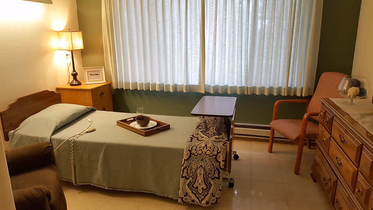 A small, cozy bedroom in a senior living facility with a single bed covered in a green bedspread. A wooden nightstand with a lamp and a framed 'WELCOME' sign sits beside the bed. There is a patterned bench at the foot of the bed with a small overbed table. A pink cushioned chair and a wooden dresser with decorative items are also visible. The room has a large window with white curtains.