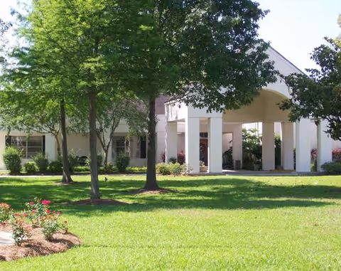 Exterior view of Capitol House Nursing & Rehab Center showing a green lawn with trees and flower beds in front of a building entrance with white pillars and a covered driveway.