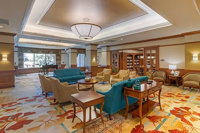 A spacious and well-lit living room area in a senior living facility featuring comfortable seating including teal and beige armchairs and sofas arranged around wooden coffee tables. The room has a colorful patterned carpet, large windows letting in natural light, and a grand piano near the window. There are built-in bookshelves and warm lighting fixtures on the walls and ceiling.