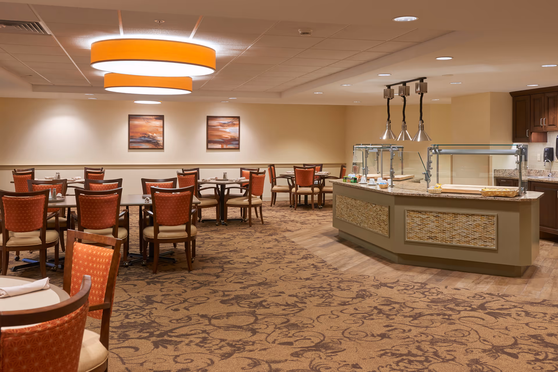 A dining room with multiple tables and chairs arranged neatly. The chairs have red upholstery with a subtle pattern. There is a serving counter with glass sneeze guards and heat lamps on the right side. The room has warm lighting with two large circular ceiling lights and two framed paintings on the far wall. The carpet has a floral pattern in shades of brown.
