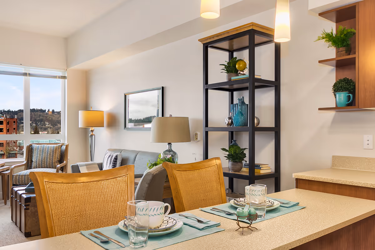 Interior view of a senior living facility showing a dining counter set for two with plates, cups, glasses, and utensils. Behind the counter, there is a living area with a sofa, armchair, floor lamp, and a black open shelving unit decorated with plants and vases. A large window reveals an outdoor view with trees and buildings.