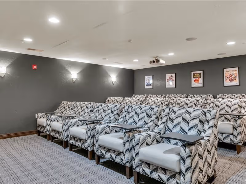 A small theater room with multiple rows of patterned armchairs equipped with individual foldable tray tables, gray walls with framed movie posters, ceiling lights, and a projector mounted on the ceiling.