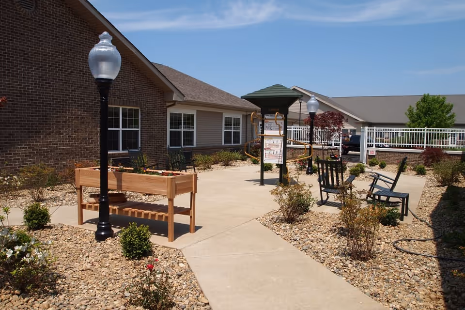 Sunny outdoor courtyard with benches, raised planters, lamp posts, and a paved walkway beside brick buildings.