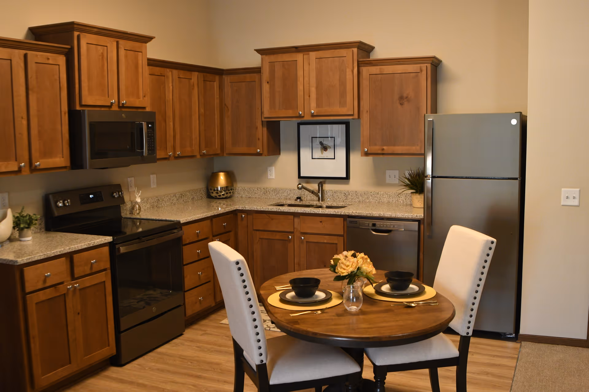 A kitchen with wooden cabinets, a stainless steel refrigerator, microwave, stove, and dishwasher. There is a round wooden dining table set for two with beige upholstered chairs, black bowls, plates, gold utensils, and a small flower arrangement in the center. The walls are beige and there is a framed picture above the sink.