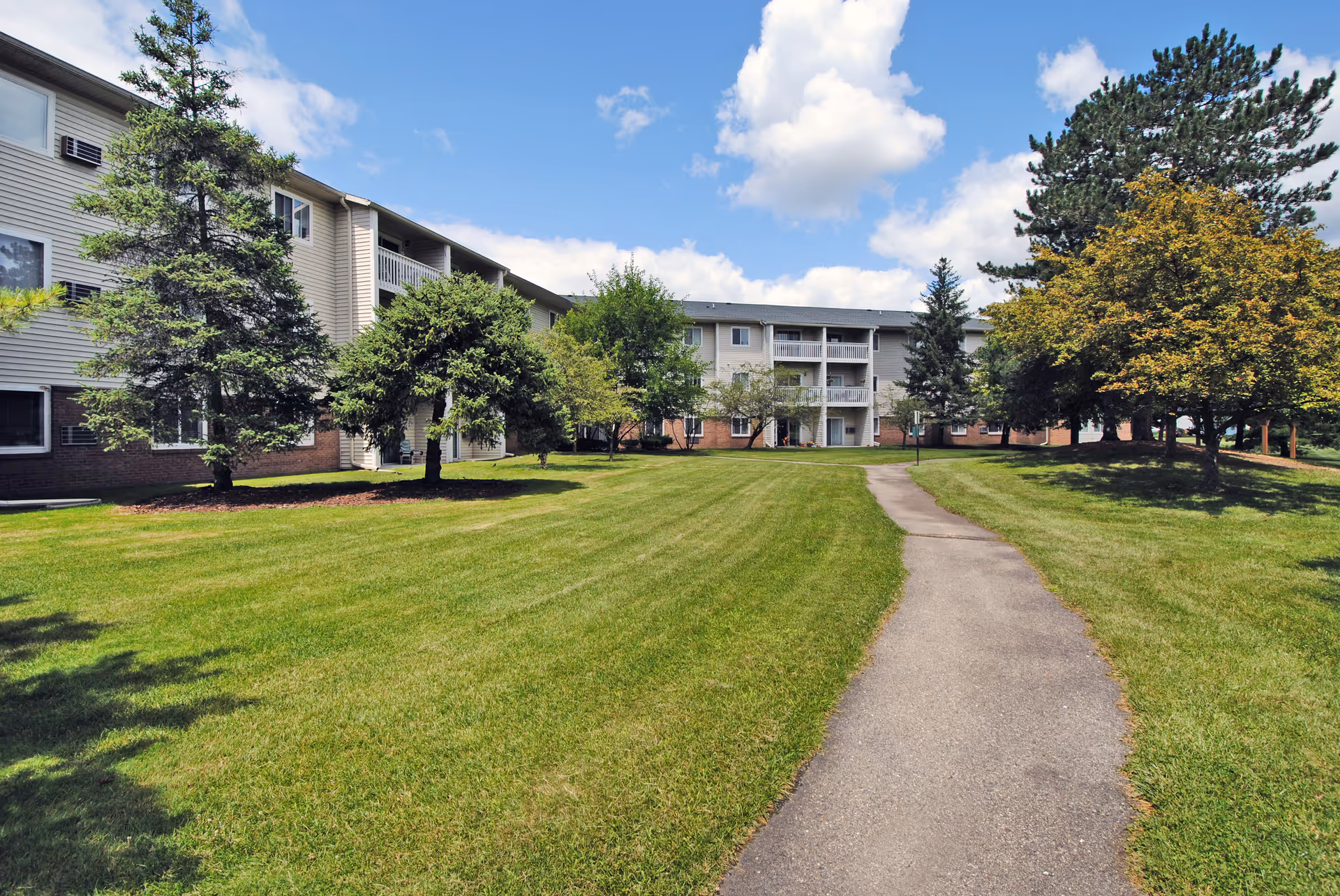 A paved walkway leads through a well-maintained grassy lawn with several trees, towards a three-story residential building with balconies under a partly cloudy blue sky.