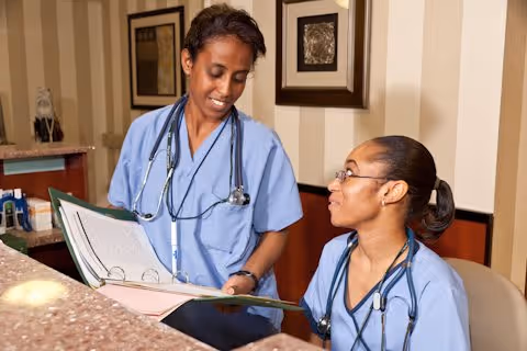 Two female healthcare professionals in blue scrubs with stethoscopes around their necks are reviewing a large binder together in an indoor setting with framed artwork on the wall behind them.