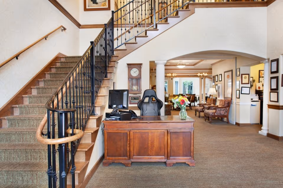 Interior view of a senior living facility reception area with a wooden desk, a computer, and a black office chair. To the left is a carpeted staircase with a wooden handrail and black balusters. In the background, there is a seating area with upholstered chairs, a dining area with tables and chairs, and framed certificates on the walls.