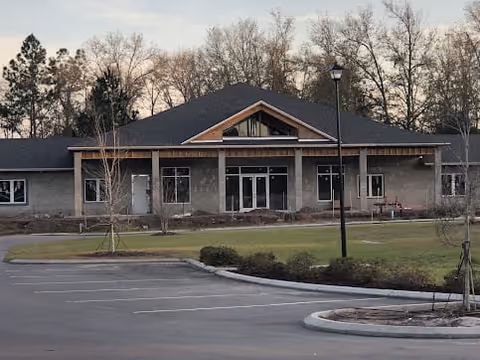 Exterior view of a single-story building with a dark roof and multiple windows, surrounded by a parking lot and landscaped greenery with trees in the background during dusk.