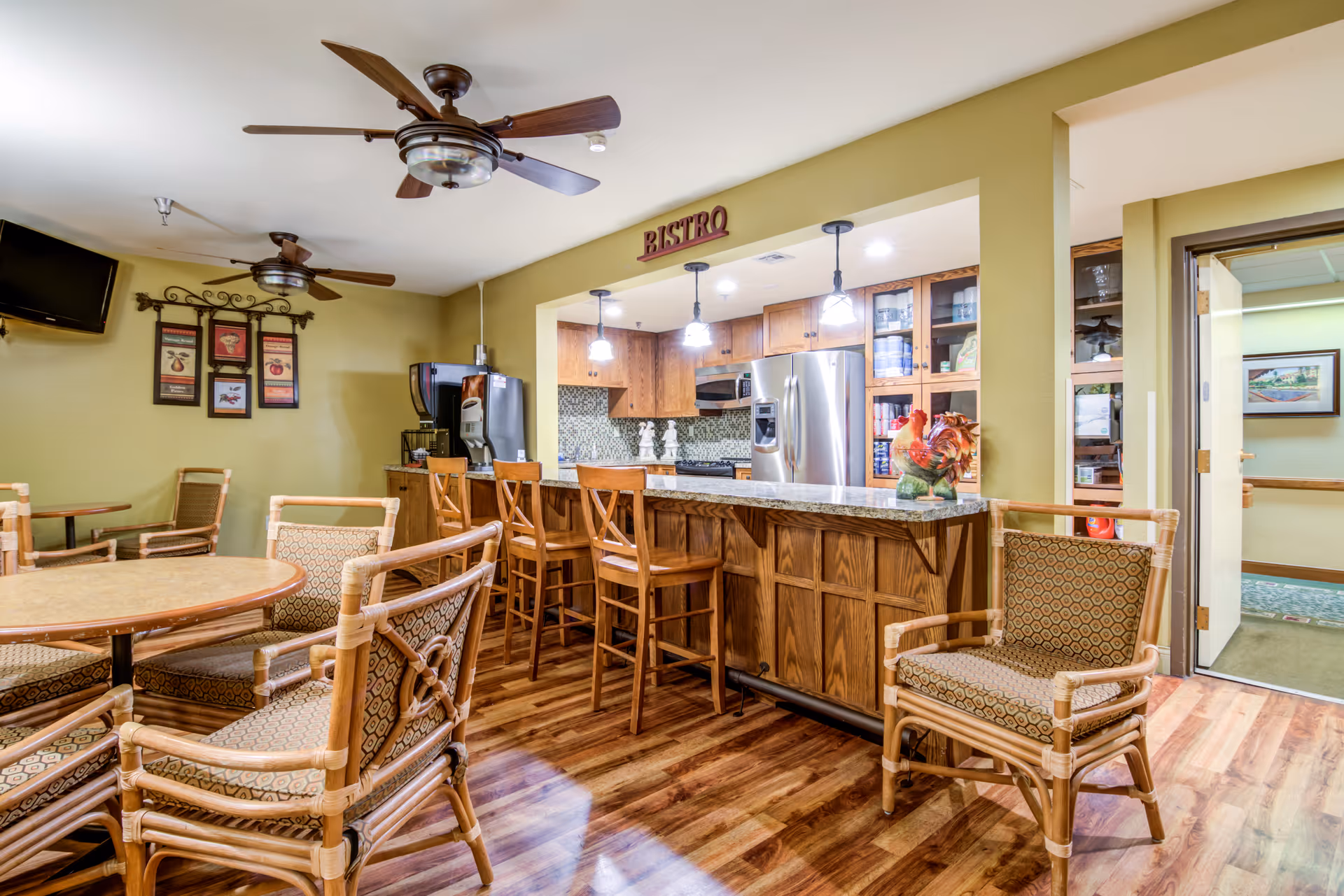 Interior view of a senior living facility's bistro area featuring a wooden bar counter with four high chairs, a kitchen area with stainless steel refrigerator and wooden cabinets, round tables with cushioned chairs, ceiling fans, and a wall-mounted TV. The room has warm lighting and wood flooring.