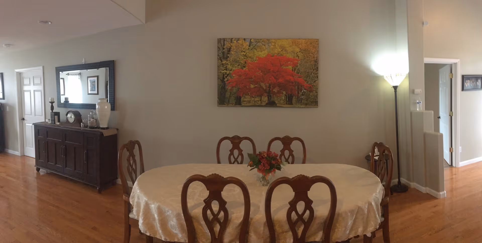 Dining room with an oval table covered with a white tablecloth and six wooden chairs around it. A small vase with flowers is placed in the center of the table. On the wall behind the table, there is a painting of a tree with red leaves. To the left, there is a dark wooden sideboard with decorative items including a large white vase, a clock, and candlesticks. A large mirror hangs above the sideboard. The room has wooden flooring and light-colored walls. A floor lamp is positioned in the corner to the right, and there are doorways visible on both sides of the room.