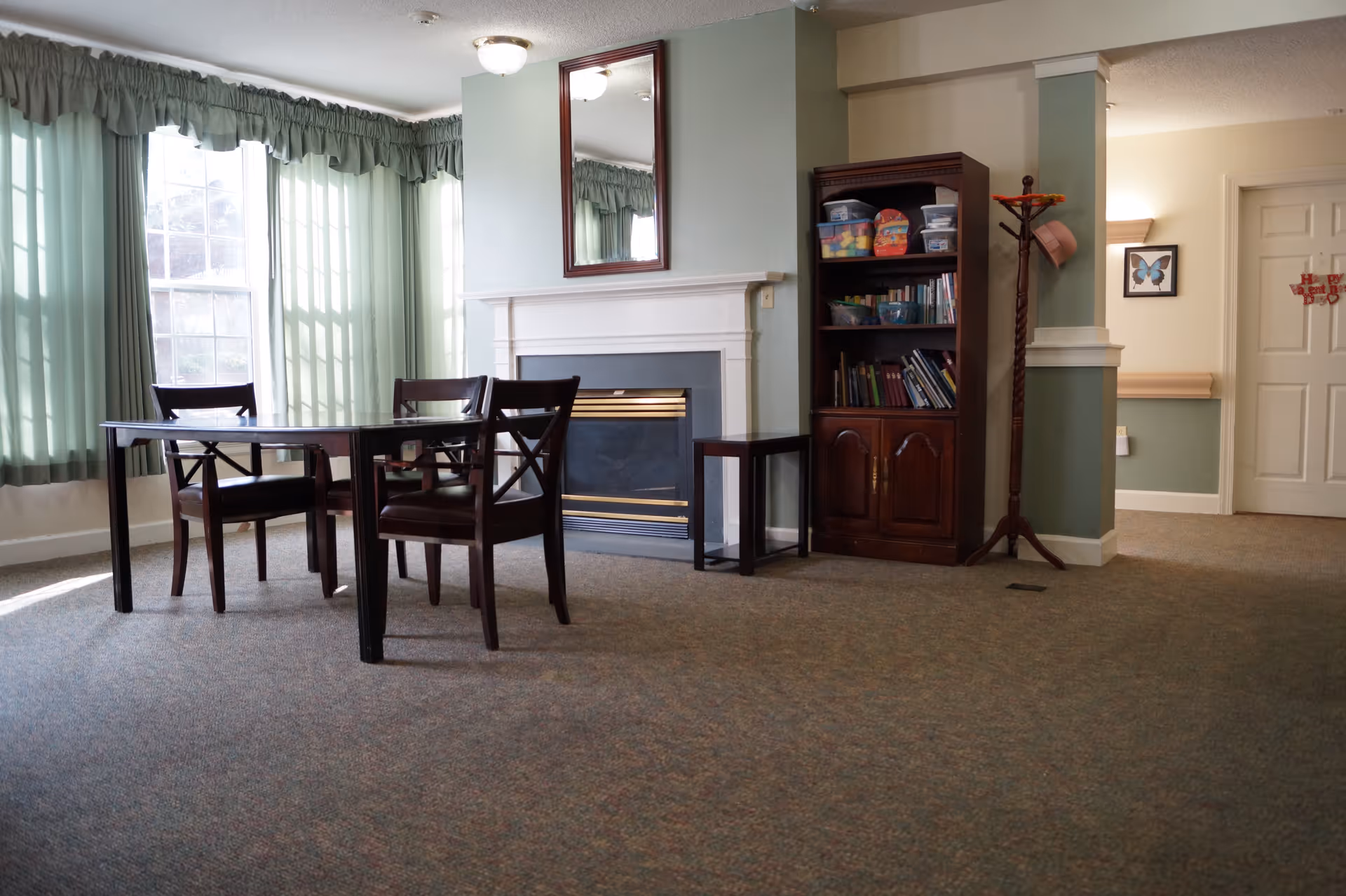 A cozy interior room with a table and four wooden chairs near large windows with green curtains. There is a fireplace with a mirror above it, a wooden bookshelf filled with books and containers, and a coat rack with a hat. The room has carpeted flooring and soft lighting.