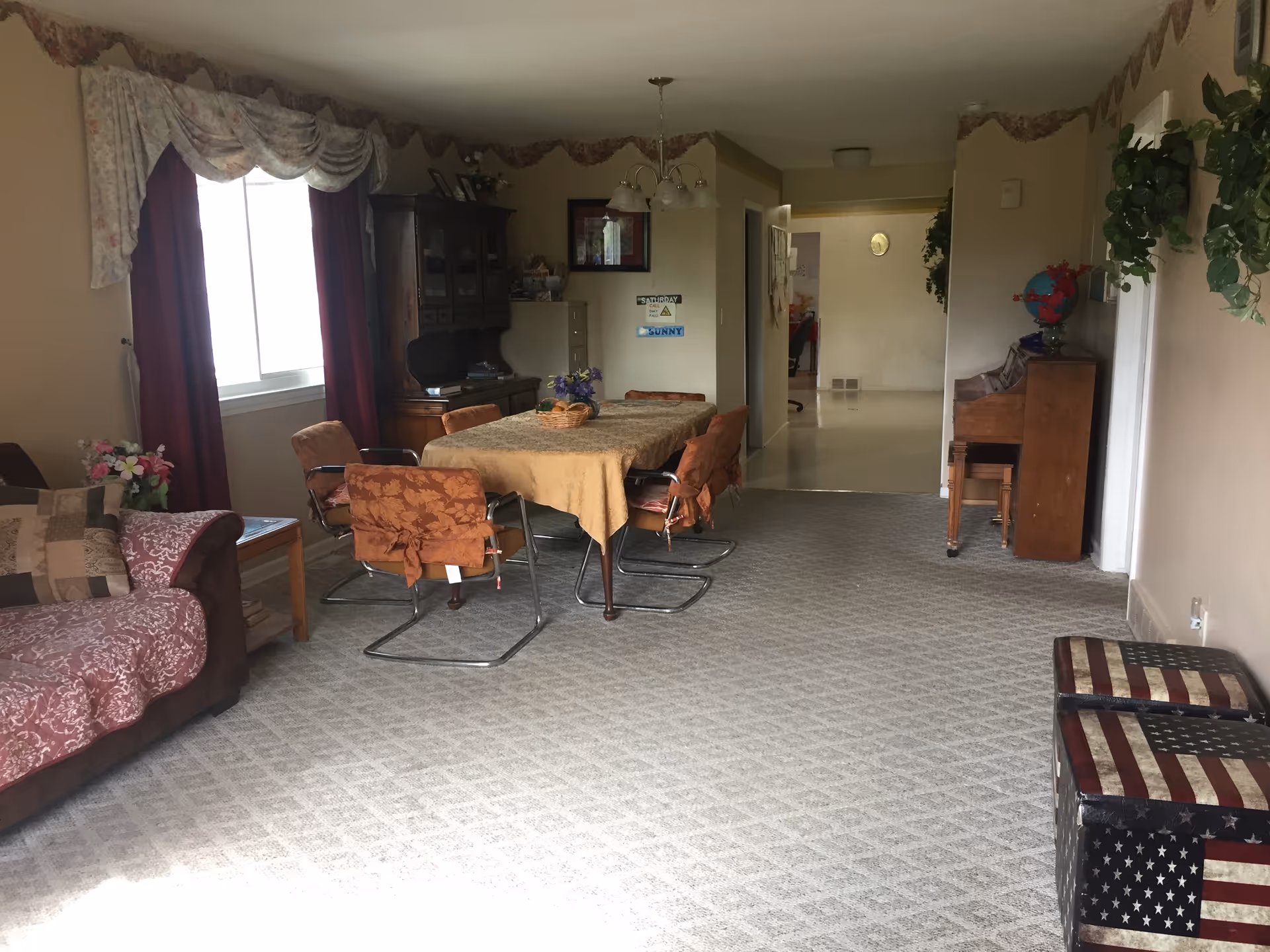 Carpeted communal living and dining area with a table and chairs, a sofa, a piano, and window with curtains.