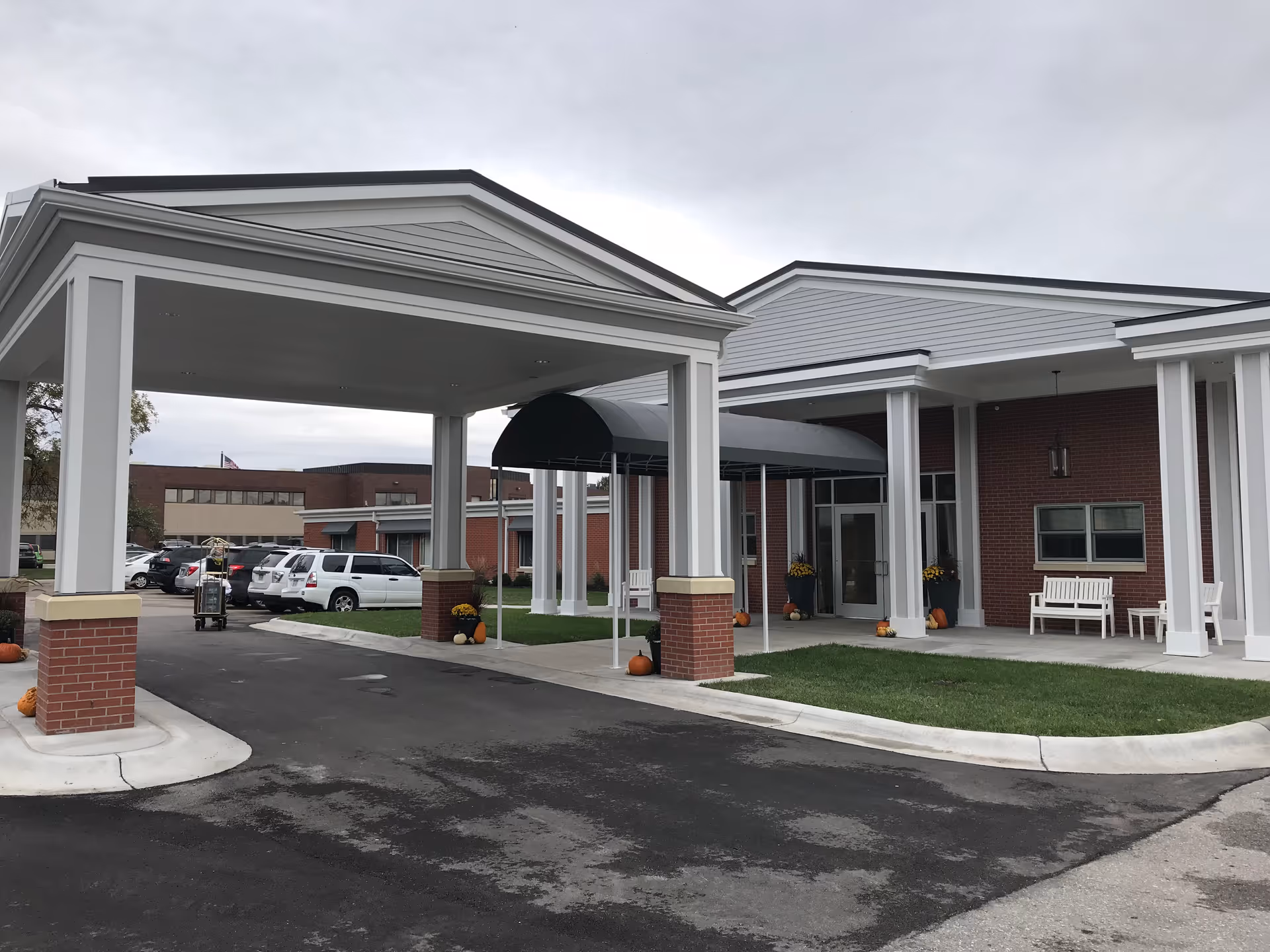 Entrance of a senior living facility with a covered driveway supported by white columns with brick bases. There are pumpkins and potted plants near the entrance, a white bench on the porch, and several parked cars in the background under a cloudy sky.