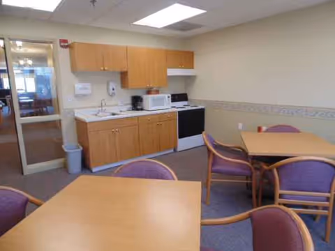 A small kitchenette with wooden cabinets, microwave, stove, and adjacent dining tables.
