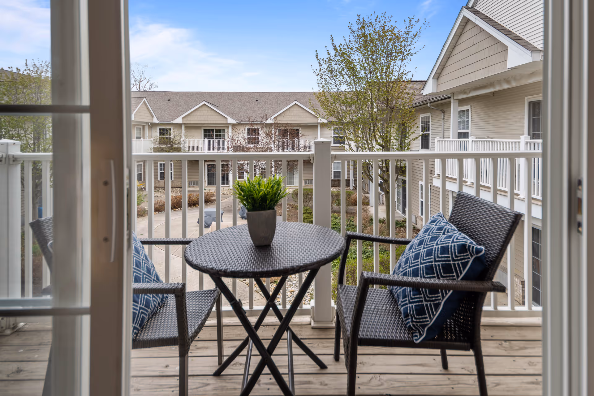 View from a balcony with a small round table and two wicker chairs with blue patterned cushions. A potted green plant is on the table. The balcony overlooks a courtyard with trees and a two-story beige building with white railings and multiple windows.