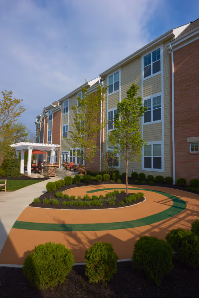 Outdoor view of a senior living facility showing a landscaped garden area with a circular walking path, small trees, and bushes. The building has three stories with beige and brick exterior walls and multiple windows. There is a white pergola with seating and a red umbrella on the patio area.
