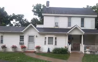 Exterior view of a two-story white house with a covered porch entrance, several windows, and flower pots along the front. There is a concrete walkway leading to the entrance and green grass surrounding the house.