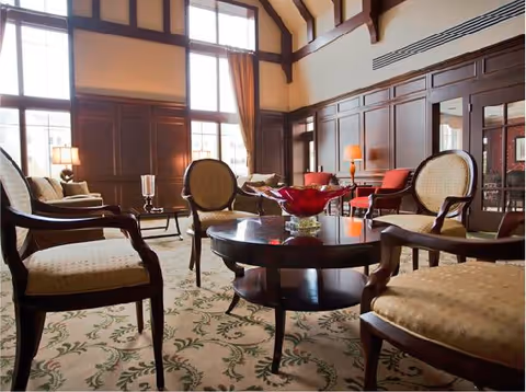 Sunlit lounge with upholstered armchairs around a round coffee table, tall windows, and wood-paneled walls.