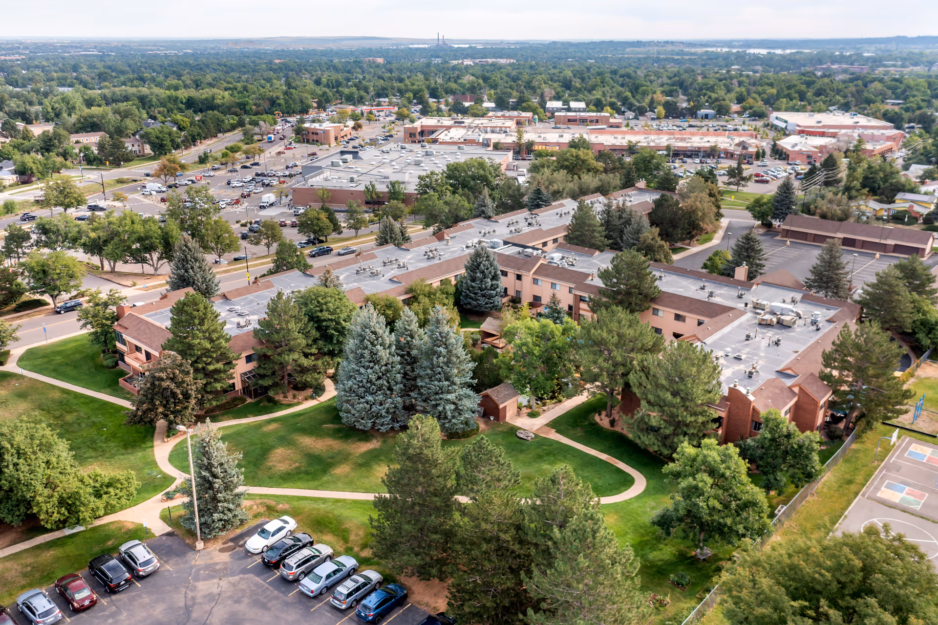 Aerial view of Brookdale Meridian Boulder facility showing multiple connected buildings surrounded by green lawns, trees, and parking lots. In the background, there is a commercial area with various buildings and parking spaces.