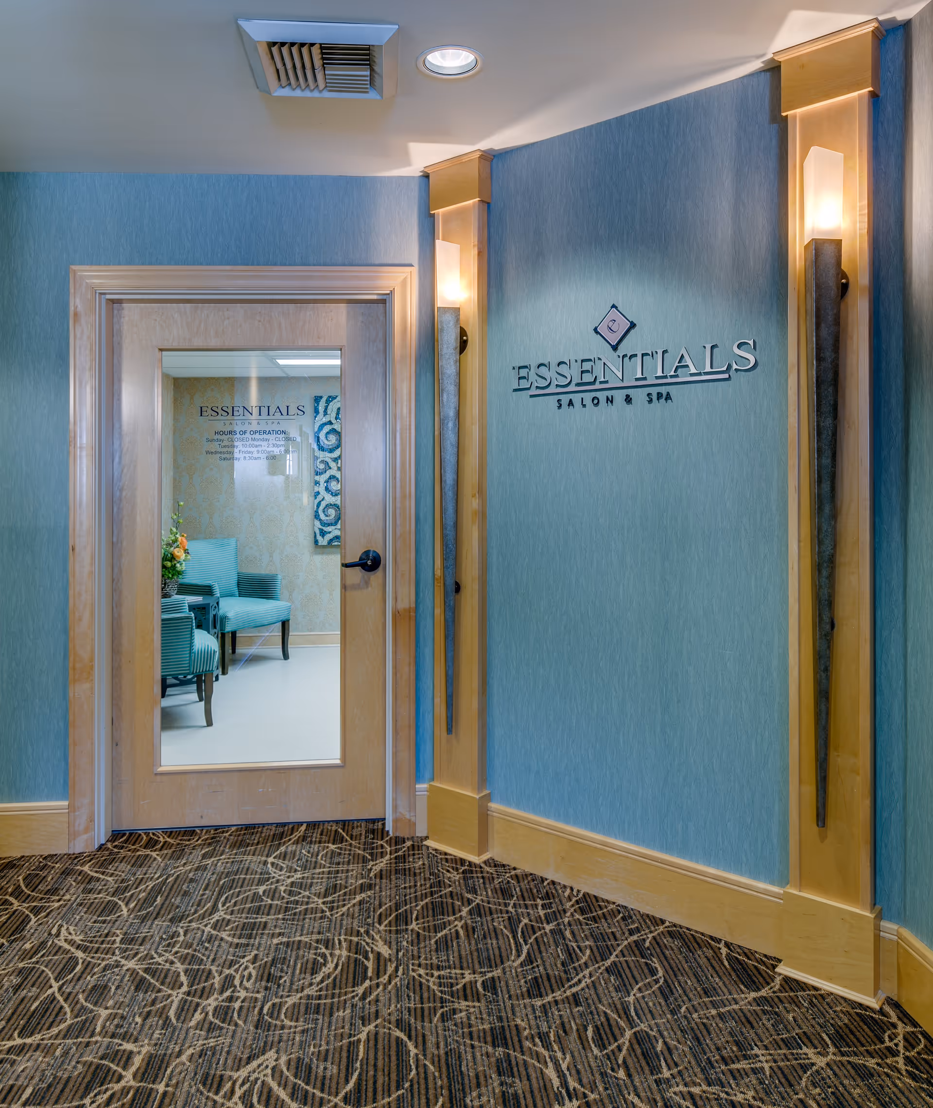 Entrance to Essentials Salon & Spa with a wooden door featuring a glass panel. Inside, there are teal upholstered chairs and a floral arrangement. The walls are blue with wooden trim and two vertical wall sconces providing light.