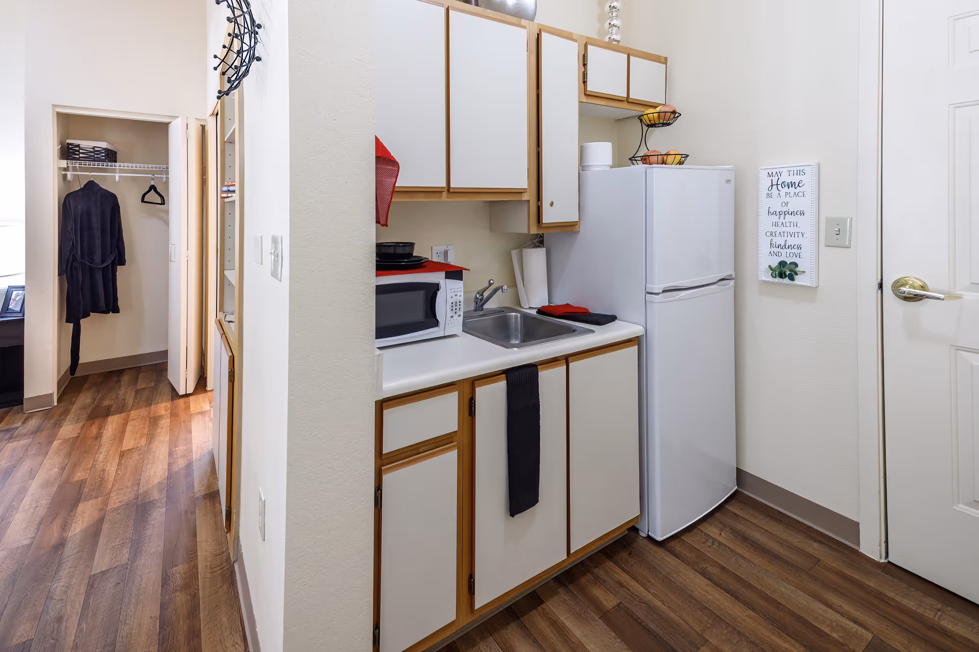 Small kitchen area with white cabinets and wooden trim, a white refrigerator, a microwave, a sink, and a two-tier fruit basket on top of the fridge. A black towel hangs from a cabinet handle. To the left, there is an open closet with a black robe hanging inside. The floor is wood-patterned, and a decorative sign is mounted on the wall next to the refrigerator.
