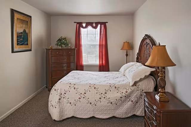A bedroom with a bed covered in a white quilt with floral patterns, two pillows, and a wooden headboard. There is a wooden nightstand with a lamp on the right side of the bed, a tall wooden dresser with a plant on top against the left wall, a window with red curtains in the center, and a framed painting on the left wall.