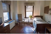 A bright single-occupancy bedroom with a bed by two windows, a chair, small table with a plant, dresser and TV.