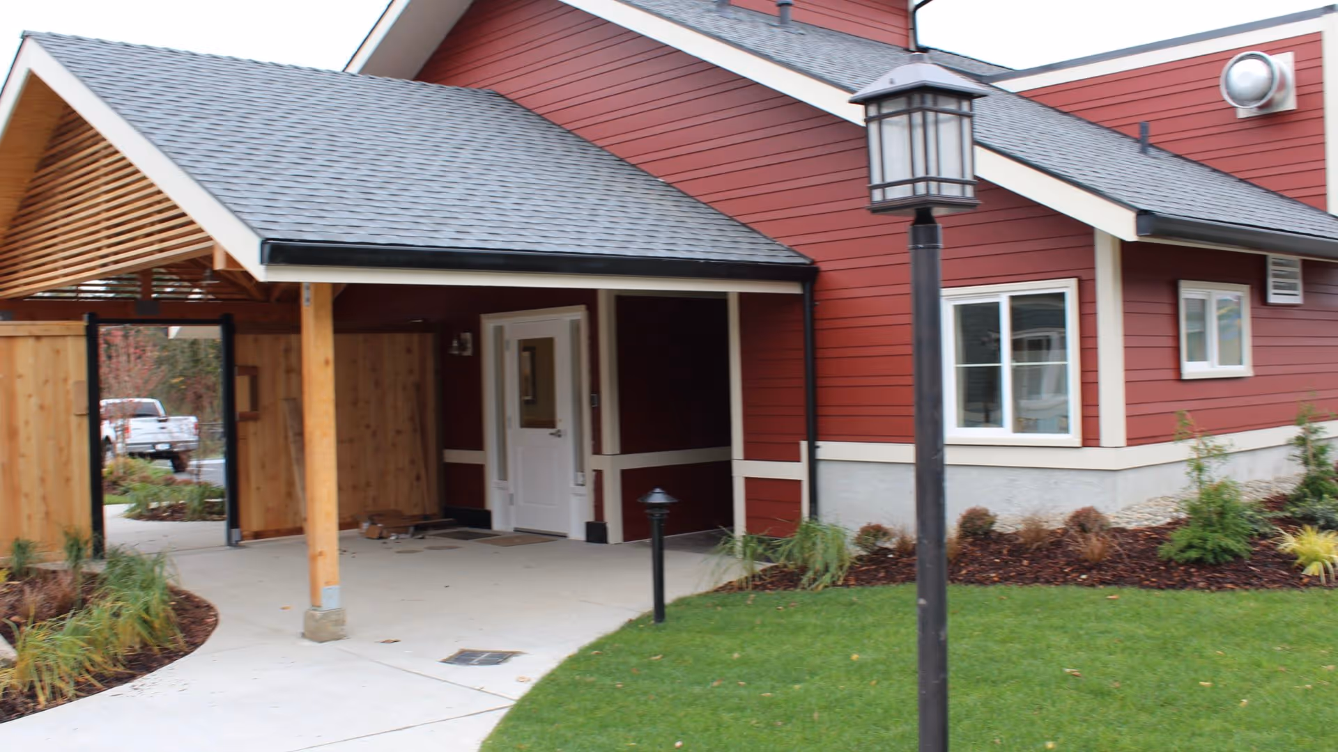 Exterior view of a red building with white trim, featuring a covered entrance supported by wooden posts. There is a concrete walkway leading to a white door, landscaped greenery, and a black lamp post on a grassy area in front of the building.