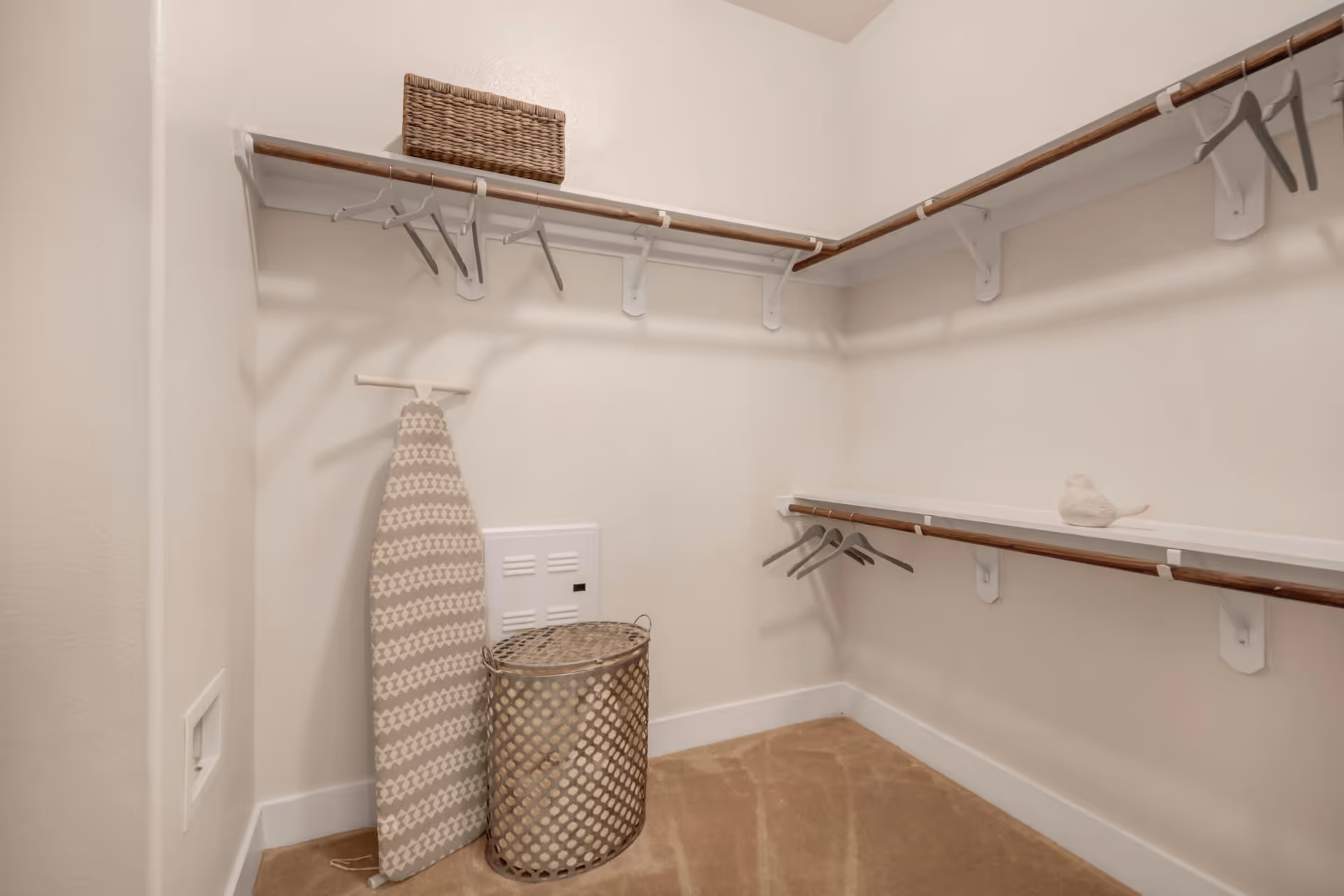 A walk-in closet with beige carpet, white walls, and wooden hanging rods with white brackets. There are several empty hangers on the rods, a patterned ironing board leaning against the wall, a woven laundry basket, a small decorative bird on a shelf, and a wicker basket on the top shelf.