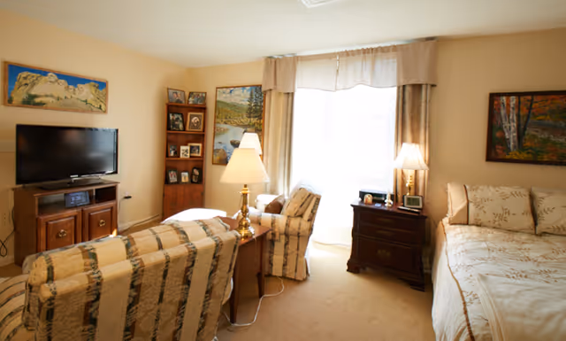 Sunny assisted-living studio room with a bed, two upholstered chairs, a TV, side tables and lamps, and framed artwork on the walls.