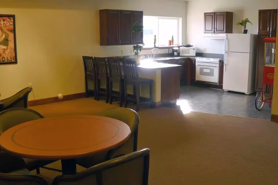 Communal lounge area with a round dining table and chairs in the foreground and an open kitchen with island, bar stools, refrigerator and stove in the background.