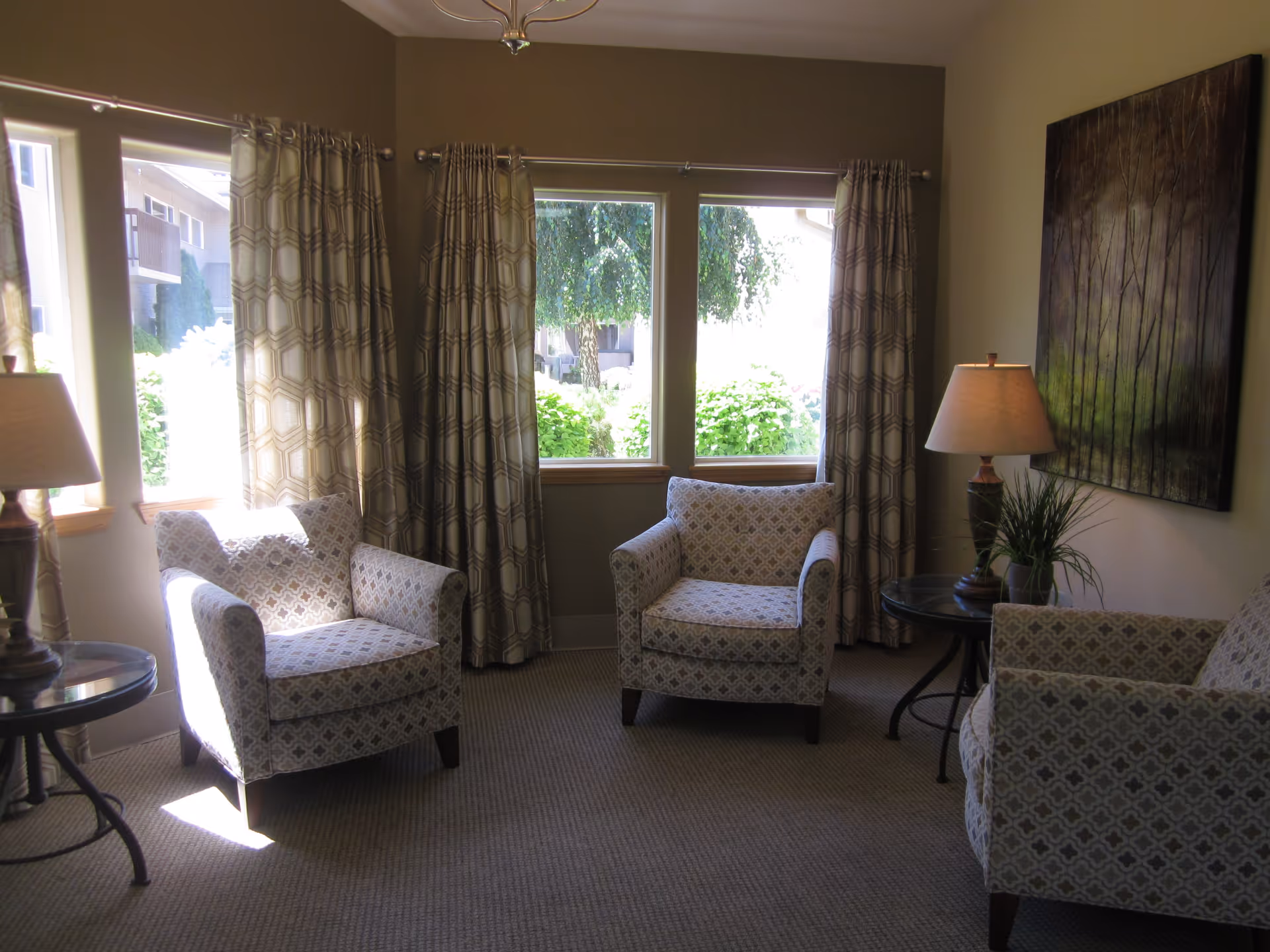 A cozy living room area with three patterned armchairs arranged around two small round side tables, each with a lamp. The room has large windows with patterned curtains allowing natural light to brighten the space. A large painting with a nature theme hangs on the wall.