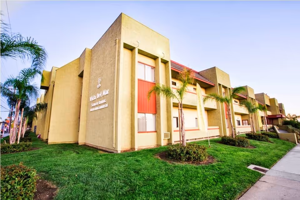 Exterior view of Vista Del Mar Senior Living building with beige walls, red accents, and several palm trees planted along the green lawn. The building has multiple windows and a sidewalk running alongside it.