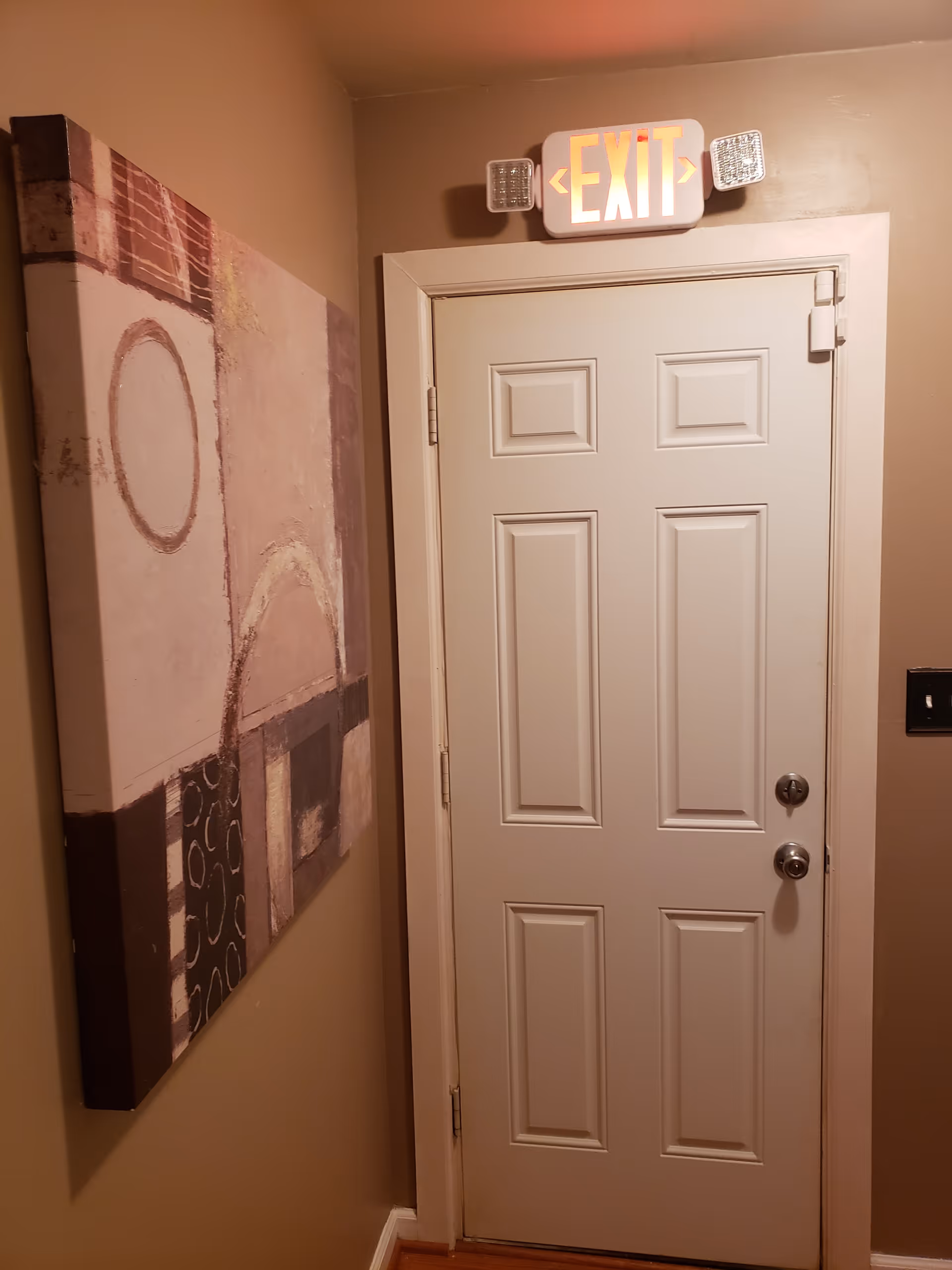 Interior view of a beige hallway corner with a white door featuring two locks and an illuminated exit sign above it. To the left of the door, there is a large abstract painting with circular and rectangular shapes in muted colors.