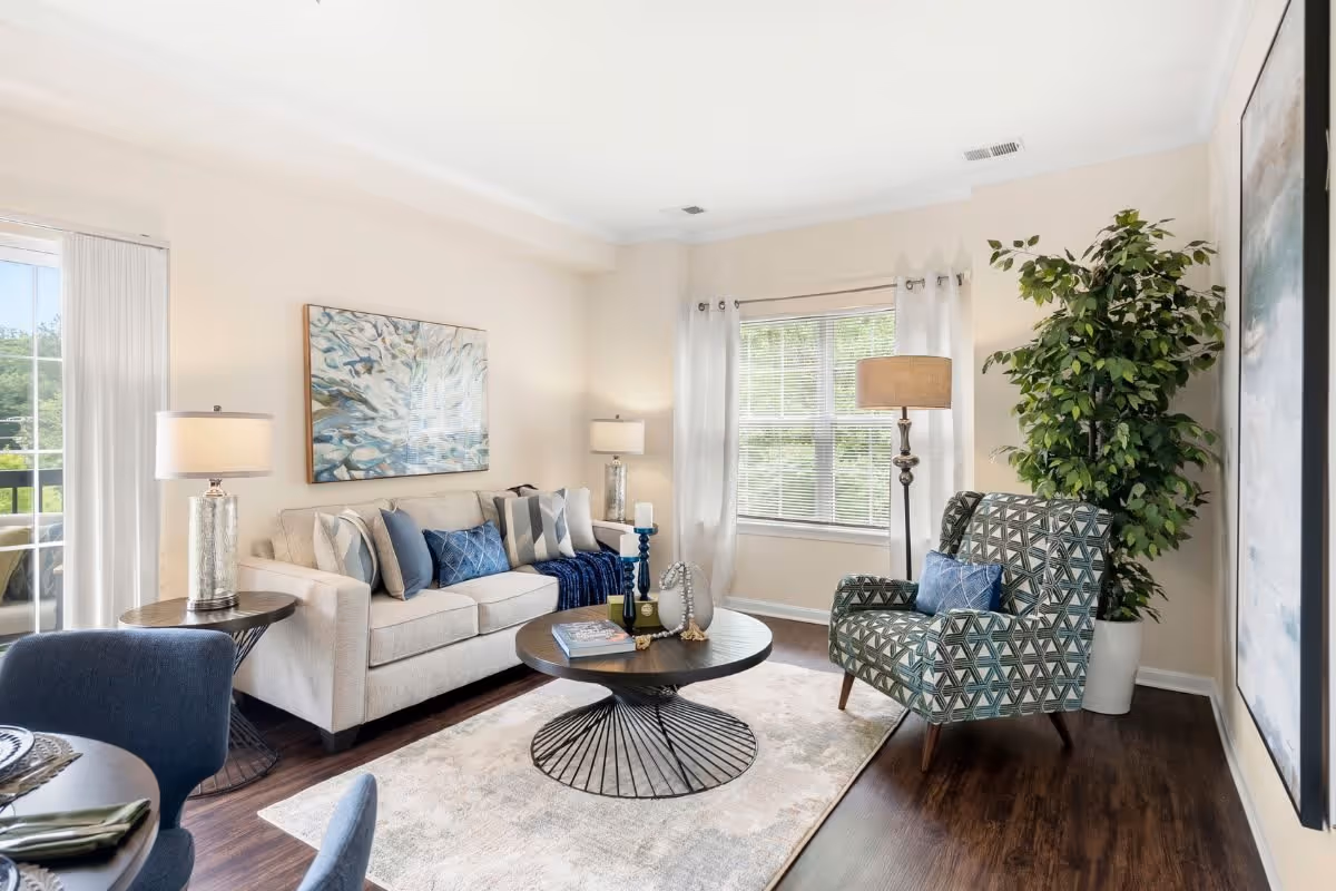 A bright and cozy living room with a beige sofa adorned with multiple cushions, a patterned armchair with a blue pillow, a round coffee table with decorative items, two table lamps, a floor lamp, a large potted plant, and a large window with white curtains letting in natural light. A piece of abstract artwork hangs above the sofa.