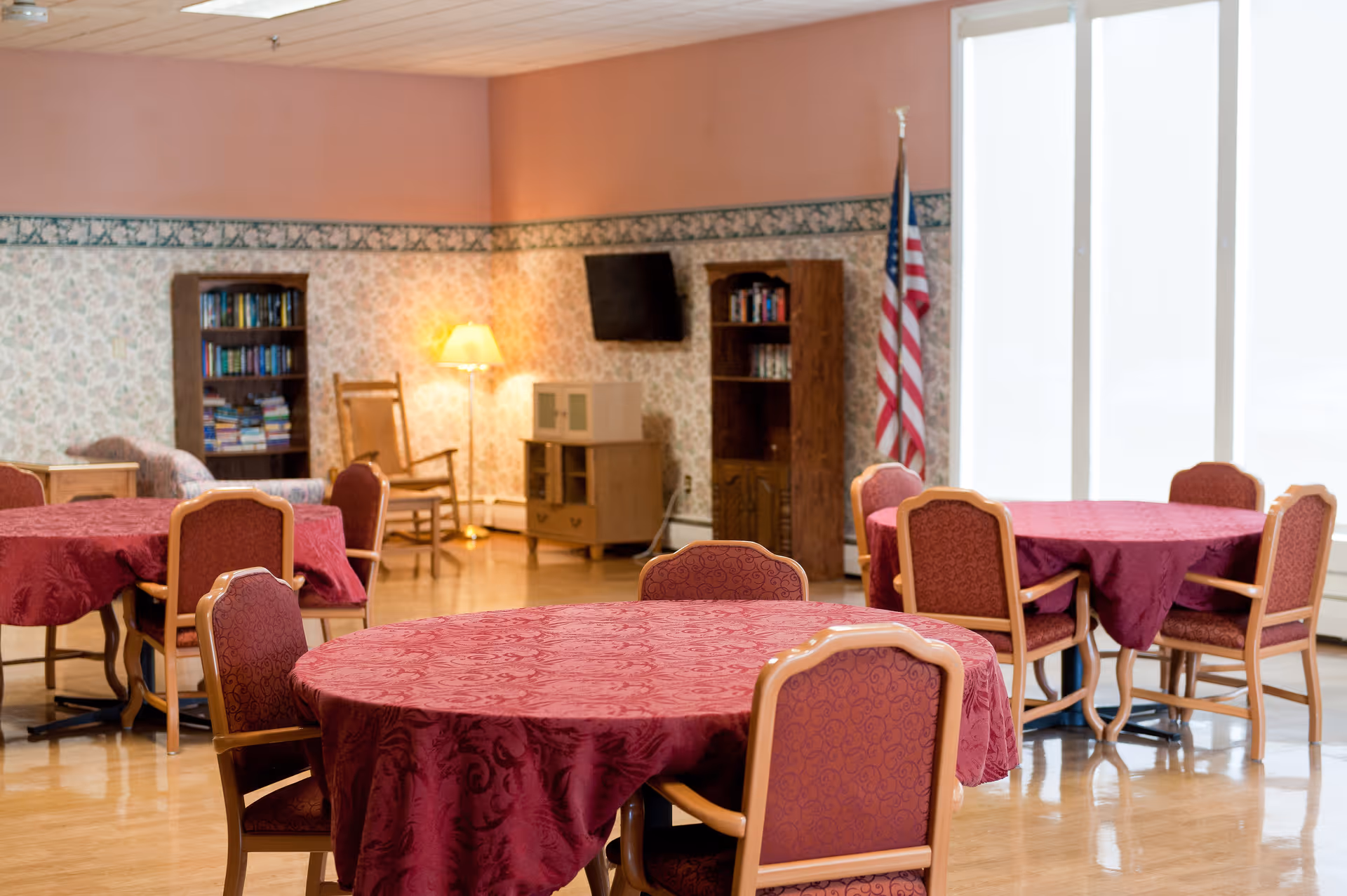 A common area in a senior living facility with round tables covered in red tablecloths surrounded by chairs. The room has floral wallpaper with a border, a wooden rocking chair, bookshelves, a floor lamp, a TV mounted on the wall, and an American flag near large windows with white blinds.