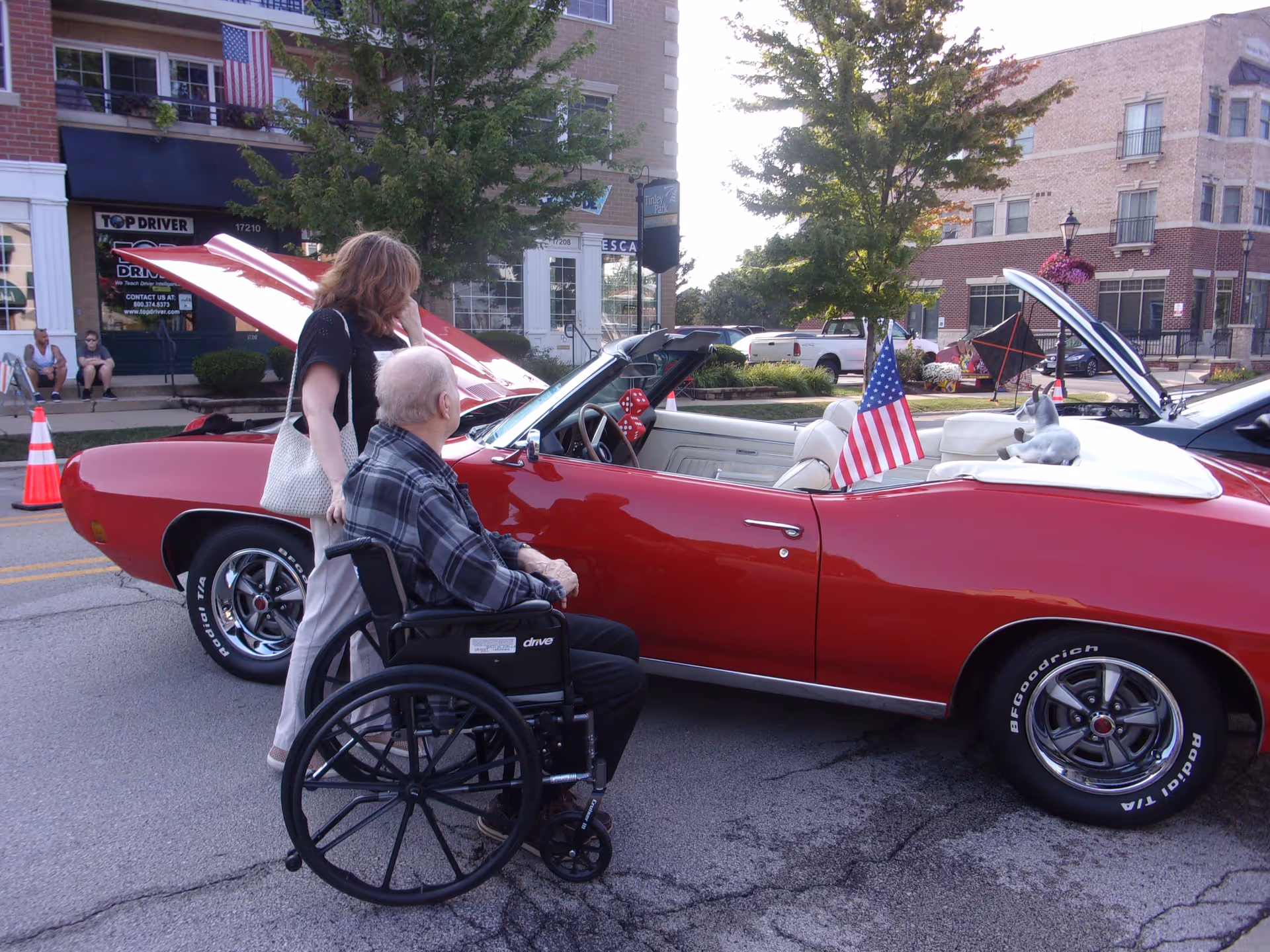 An elderly man in a wheelchair and a woman standing beside him are looking at a red classic convertible car with its hood open, parked on a street in a small town setting. The car has an American flag attached to it and a small dog sitting on the back seat. There are buildings, trees, and other parked cars in the background.