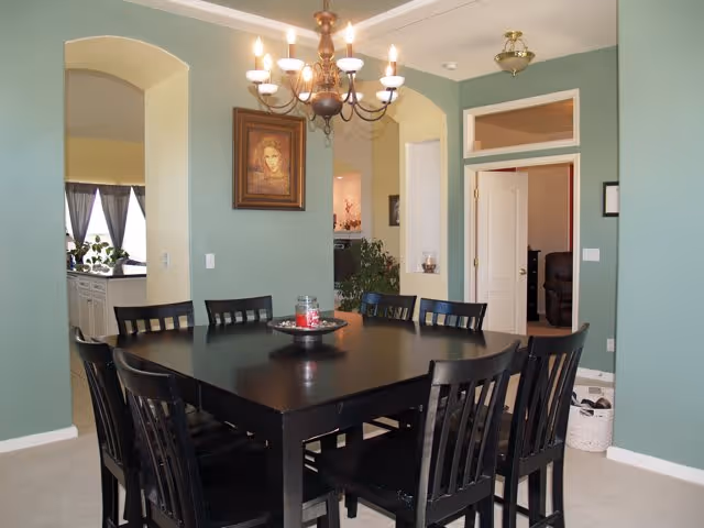 Dining room with a black rectangular table surrounded by eight black chairs. A chandelier with lit bulbs hangs above the table. The walls are painted green with white trim, and there is a framed portrait on the wall. An open doorway leads to another room, and a window with curtains is visible in the background.
