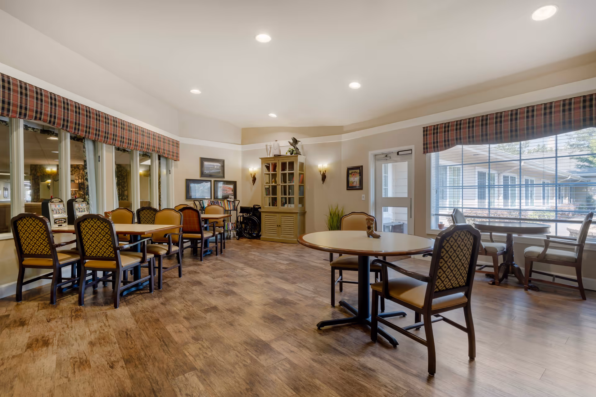 A bright dining area in Cinco Ranch Alzheimer's Special Care Center with multiple tables and chairs arranged neatly. The room has large windows with plaid valances, allowing natural light to fill the space. There is a cabinet with decorative items and framed pictures on the walls. The floor has a wood-like finish, and the overall atmosphere is clean and welcoming.