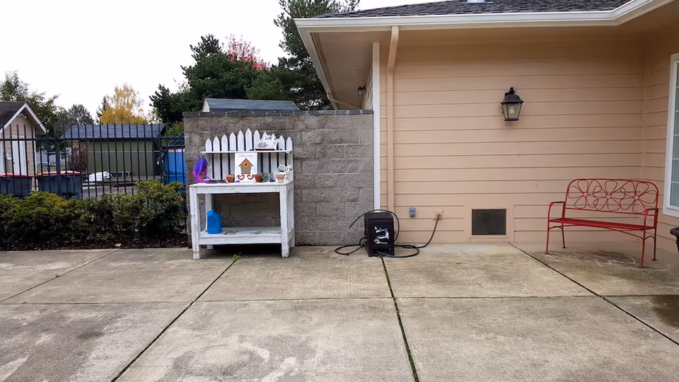 Outdoor patio area with concrete flooring, a beige building wall with a black lantern light fixture, a red metal bench with floral design, a white wooden shelf with various small items including a birdhouse, and a black hose reel on the ground. There are bushes and a black metal fence in the background.