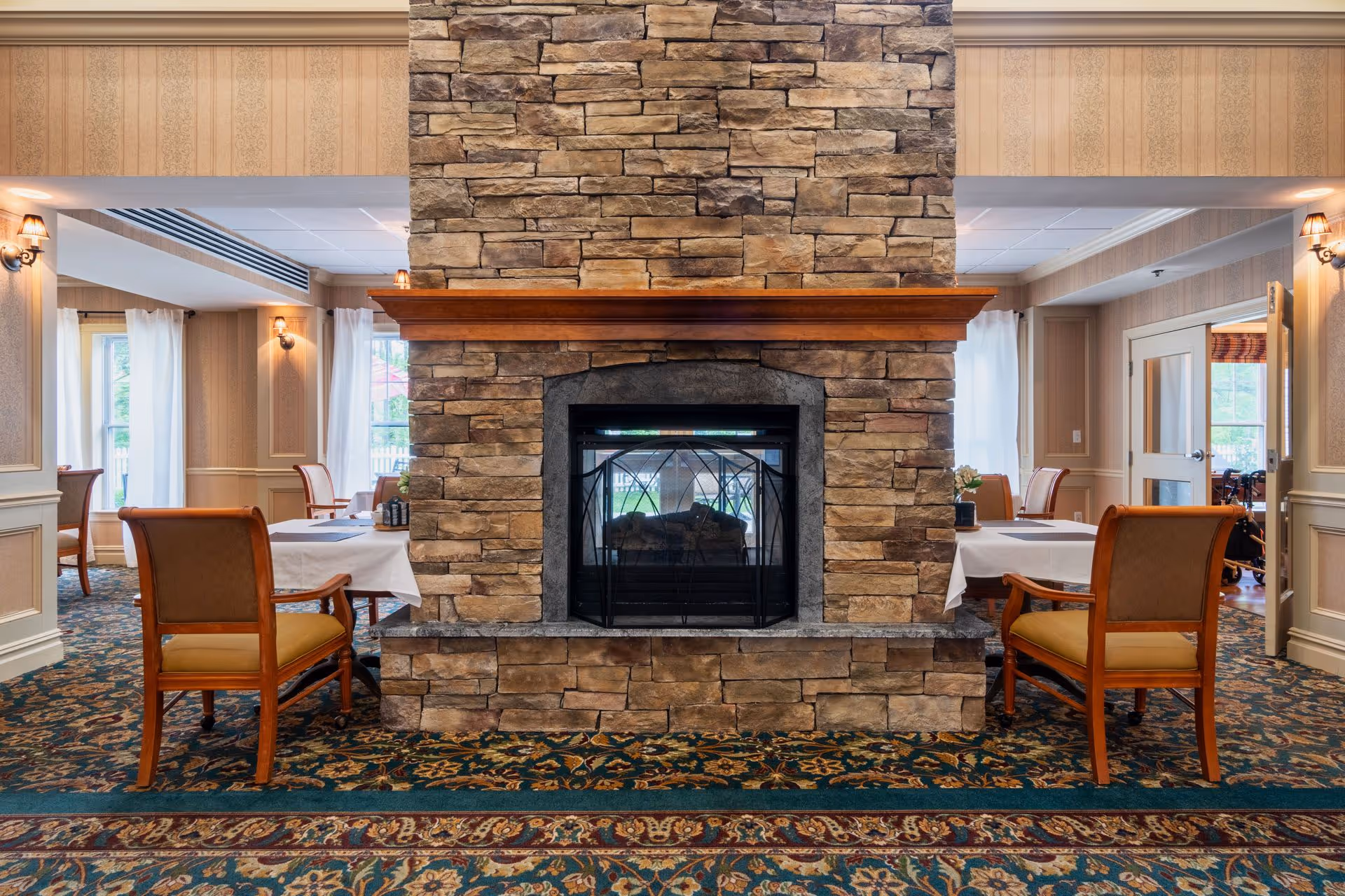 A cozy dining area with a large stone fireplace in the center. The room features patterned carpet, wallpapered walls, and wooden chairs with cushioned seats around tables covered with white tablecloths. Natural light comes through windows with white curtains.